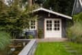 Garden with a small dark wooden shed, white doors, stone path, and pond surrounded by lush greenery.