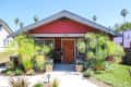Red bungalow with a wooden door, surrounded by lush plants and a small front yard.
