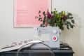Sewing machine on black table with floral arrangement, clear chair, and pink "For Like Ever" poster on wall.