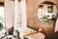 Bathroom with a white tiled shower, round mirror, and plants, reflecting a cozy living room with a sofa and large window.