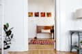 Hallway view into a room with a wooden bench, red striped rug, and four colorful paintings on the wall.