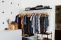 Open closet with hanging clothes, white dresser, shoe rack, and a small cart in a room with cross-patterned wall.