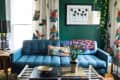Blue tufted sofa with colorful cushions, floral curtains, and a vase of purple flowers on a coffee table in a green-walled room.