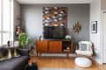 Mid-century modern living room with a TV on a wooden console, patterned wall art, guitar, and a cozy chair with a pouf.