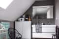 Bathroom with sloped ceiling, large mirror, double sink, white cabinets, black chair, and patterned towel.