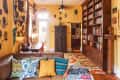 Living room with yellow walls, tribal masks, a brown leather sofa, colorful pillows, a large bookshelf, and a patterned rug.