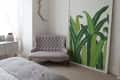 Bedroom with tufted gray loveseat, large green leaf artwork, and white dresser near a window.