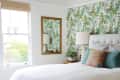 Bedroom with tropical leaf wallpaper, white bed, colorful pillows, wooden mirror, and potted plant on nightstand.