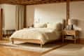 Cozy bedroom featuring a light beige bedspread, wooden bed frame, nightstand with a lamp, and a decorative plant.