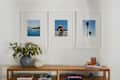 Three framed photos on a wall: a coastal view, a woman with a child at the beach, and palm trees. A vase with flowers and books on a wooden shelf.