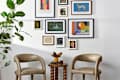 Two beige chairs beside a twisted wooden side table with a small plant and candle, against a gallery wall of framed art.