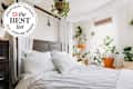 Bed with pale gray and white cotton bedding, wood headboard, white walls, sheer curtains behind bed, window letting in lots of light, multiple plants. Seal in upper left corner reads "Apartment Therapy the Best List 2023 Cooling Sheets"