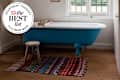 A blue freestanding bathtub with a wooden tray, a small wooden stool, and a colorful patterned rug on a woven floor.
