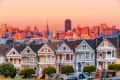 Row of colorful Victorian houses with San Francisco skyline at sunset.