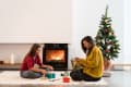 Two women wrapping gifts by a fireplace, with a decorated Christmas tree nearby.