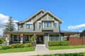 Two-story gray house with white trim, front porch, manicured lawn, and hanging pink flowers under a clear blue sky.