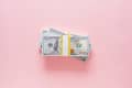 Stack of 100-dollar bills secured with a yellow band on a pink surface.