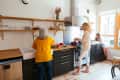 Two women cooking in a modern kitchen with open shelves, while a child sits by the window.