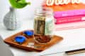 Mason jar with cash and coins on a wooden tray, next to sunglasses and a vase, with colorful books in the background.