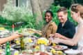 Group of friends enjoying an outdoor meal at a wooden table with flowers, wine, and salads.
