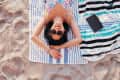 Woman in sunglasses lying on a striped beach towel, smiling on sandy beach.