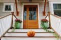 Pumpkin with carved face on porch steps, flanked by green pots with red flowers, leading to a wooden front door.