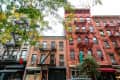 Historic brick apartment buildings with fire escapes and leafy trees in an urban setting.