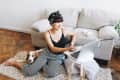 Woman in striped pants using a laptop on the floor, with a dog beside her and a gray sofa in the background.