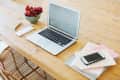 Open laptop on wooden table with a bowl of grapes, notepad, pen, and smartphone on stacked notebooks.