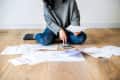 Person sitting on wooden floor, using a calculator, surrounded by scattered bills and documents.