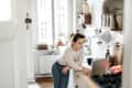 Woman in a cozy kitchen leaning on a counter, using a laptop, with kitchen utensils and plants in the background.