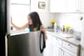 Woman opening a refrigerator in a bright kitchen with white cabinets, yellow flowers, and a cutting board on the counter.