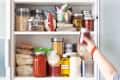 Pantry shelf with jars of grains, pasta, canned goods, and a hand reaching for a jar.