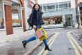 Woman in blue coat and jeans joyfully jumping on a city street, holding colorful shopping bags.