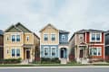 Three colorful suburban houses in a row: yellow, blue, and red, with small front yards and trees.