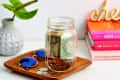 Glass jar with dollar bills and coins on a wooden tray, next to blue sunglasses, a plant, and stacked colorful books.