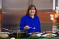 Woman in blue shirt cooking with a pan, surrounded by plates of food and flowers.