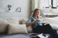 Woman in a blue shirt eating on a beige sofa, with a magazine open beside her in a bright living room.