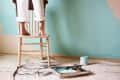 Person standing on a chair painting a wall light blue, with paint can and tray on the floor.