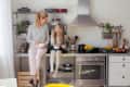 Mother and daughter eating in a modern kitchen with open shelves, fruit bowls, and stainless steel appliances.