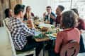 Group of friends laughing and dining together at a rustic wooden table with wine glasses and plates of food.