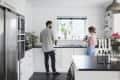 Man and woman in a modern white kitchen with black countertops, standing near a window with a potted plant.