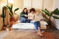 Two people sitting on a white sofa with a laptop, surrounded by potted plants in a cozy room.