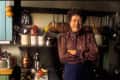 Woman in a patterned blouse standing in a kitchen with hanging copper pots, utensils, and a cutting board with ingredients.