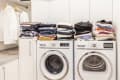 Laundry room with stacked clothes on top of a washer and dryer, white cabinets, and hanging shirts.
