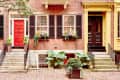 Red and black doors on a brick townhouse with window boxes, shutters, and potted plants on a cobblestone street.