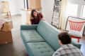 Couple moving a teal sofa in a room with moving boxes, a red chair, and a ladder.