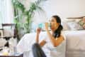 Woman sitting on floor drinking water in a cozy bedroom with plants and patterned pillows.