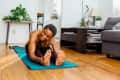 Woman stretching on a blue yoga mat in a living room with plants and a gray sofa.