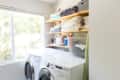 Laundry room with washer, dryer, wooden shelves, folded towels, and cleaning supplies by a large window.